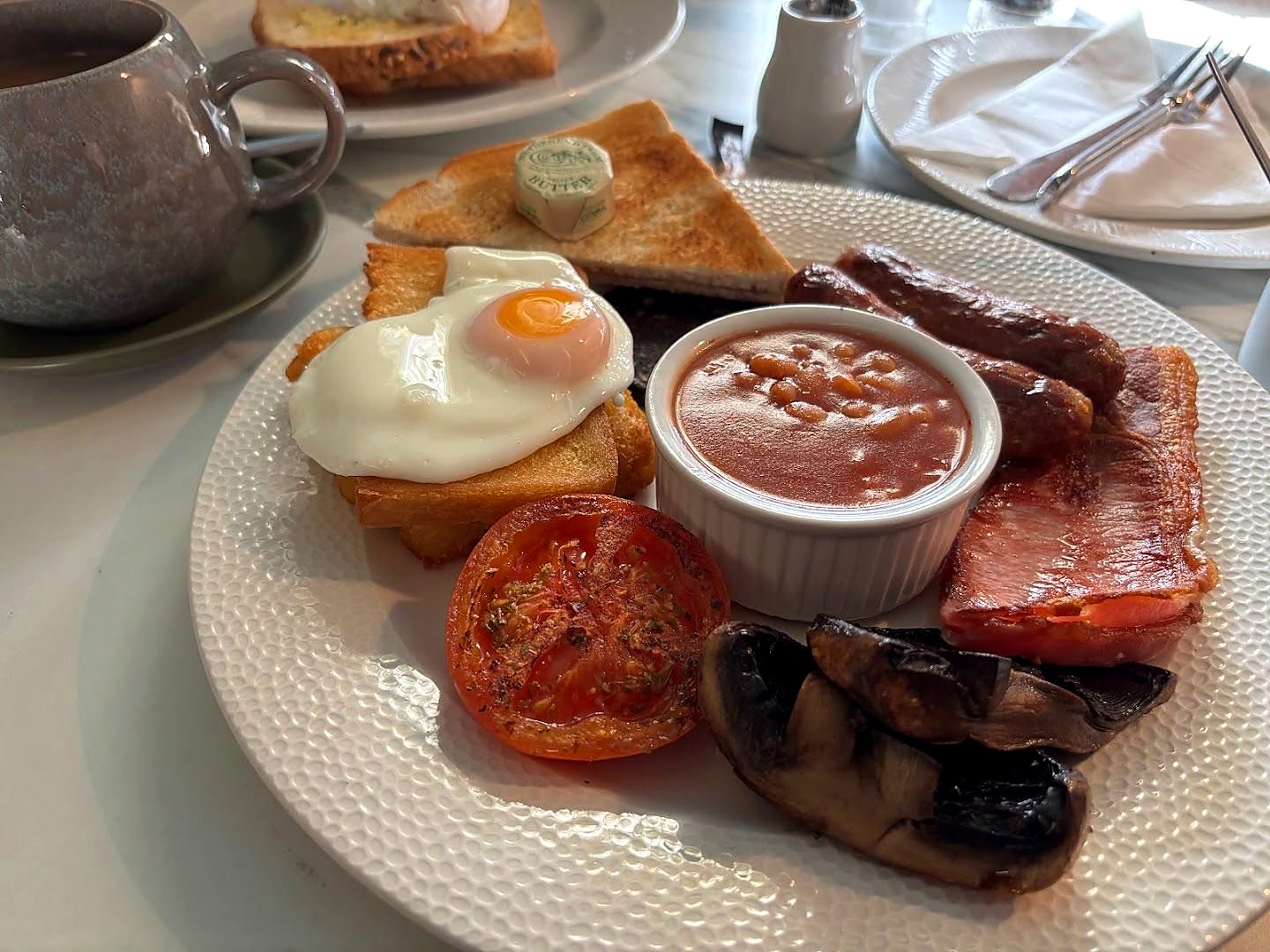 A full English breakfast on a marble table: fried egg on toast, sausages, bacon, grilled tomato, mushrooms and a pot of beans, with toast and a curl of butter