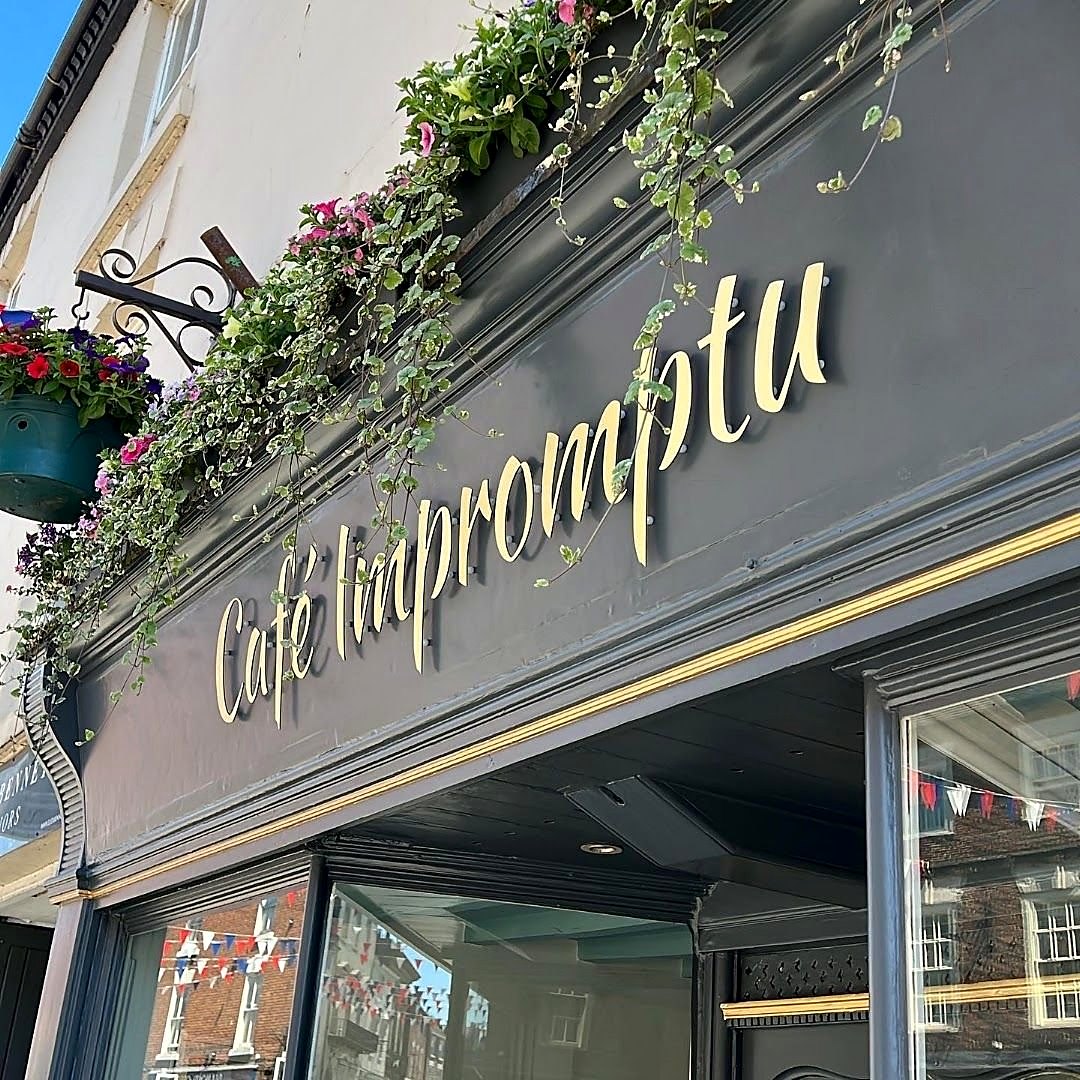 Café Impromptu’s dark grey shop sign in gold script, with flowers and ivy trailing from the planters above