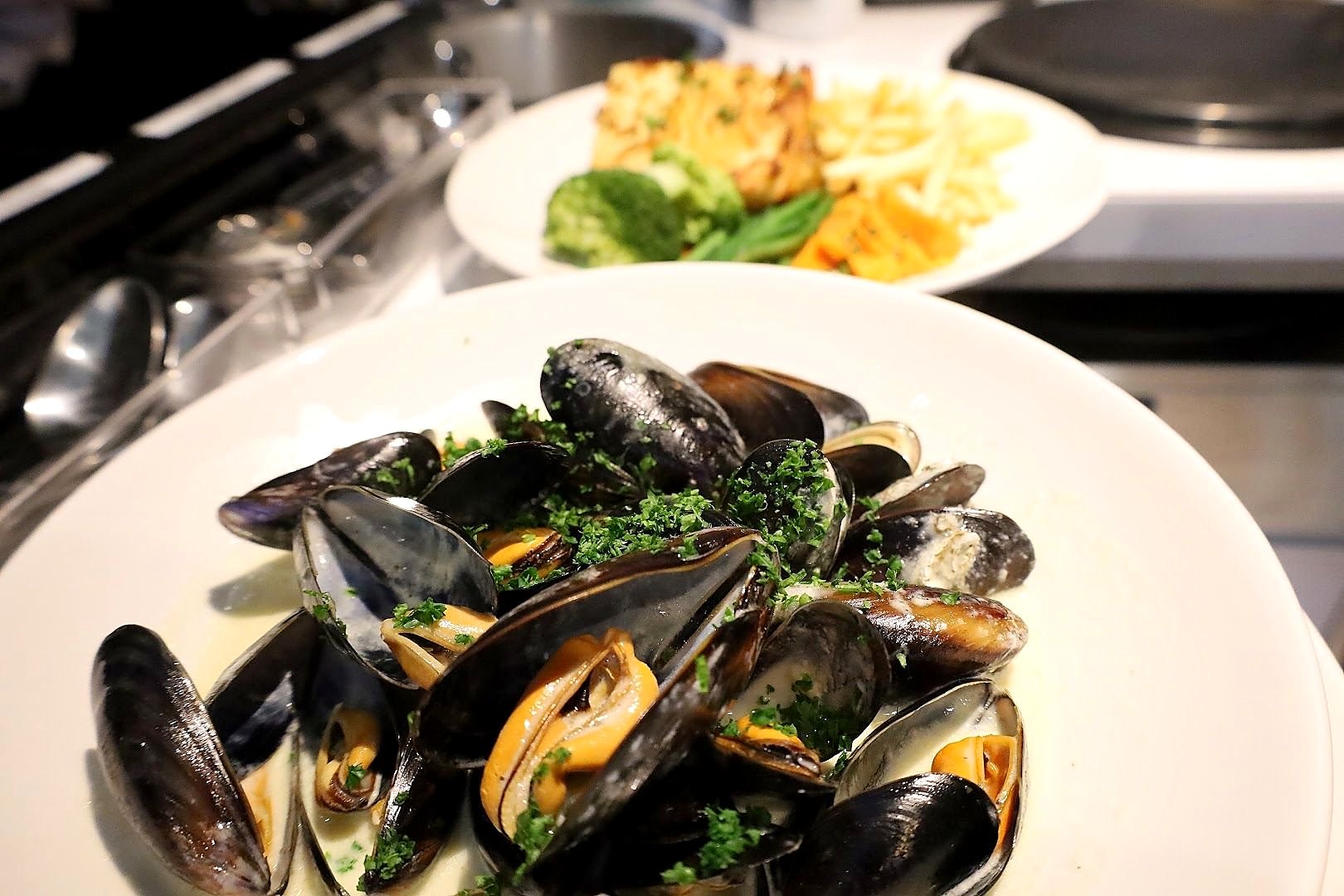 A white bowl of mussels in a creamy sauce scattered with parsley, with a plate of chips and salad behind